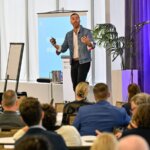 Photo taken during the Together25Ensemble conference featuring a man in a suit speaking on stage