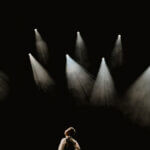 A performer on a dark stage looking up into several bright spotlights