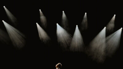 A performer on a dark stage looking up into several bright spotlights
